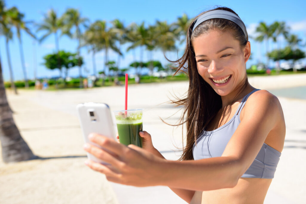 Fitness selfie woman drinking green vegetable smoothie taking self portrait photograph with smart phone after running exercise workout on beach. Diet culture with fit Asian Caucasian girl.